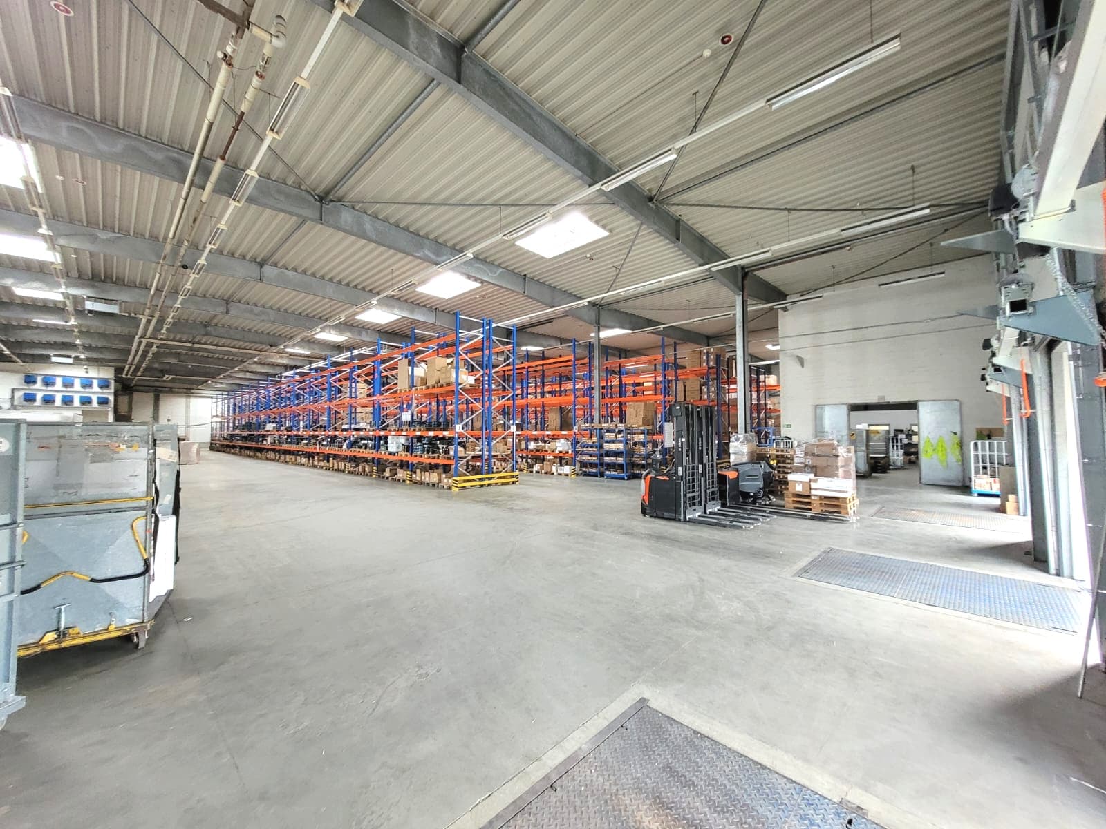 Innenansicht einer großen Lagerhalle mit hohen Regalen, Gabelstapler und Betonboden unter Metalldach.
