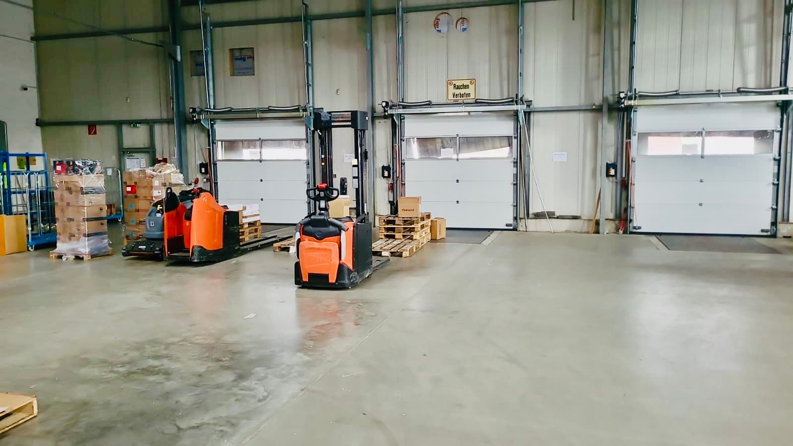 Innenansicht einer Lagerhalle mit weißen Verladetoren, orangefarbenen Gabelstaplern, Holzpaletten und verschiedenen gestapelten Kartons.